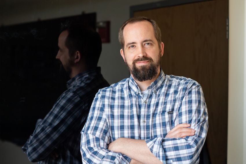 Matthew Shoulders, wearing a blue and white plaid shirt, stands with his arms crossed in front of a dark, reflective background.