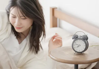 A woman getting out of bed next to an alarm clock. People with advanced sleep phase trait have early sleep onset and offset due to genetic mutations leading to shorter circadian period. A woman getting out of bed next to an alarm clock. People with advanced sleep phase trait have early sleep onset and offset due to genetic mutations leading to shorter circadian period.
