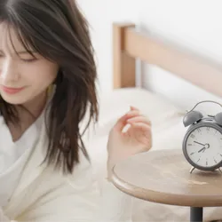 A woman getting out of bed next to an alarm clock. People with advanced sleep phase trait have early sleep onset and offset due to genetic mutations leading to shorter circadian period. 