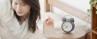 A woman getting out of bed next to an alarm clock. People with advanced sleep phase trait have early sleep onset and offset due to genetic mutations leading to shorter circadian period. 