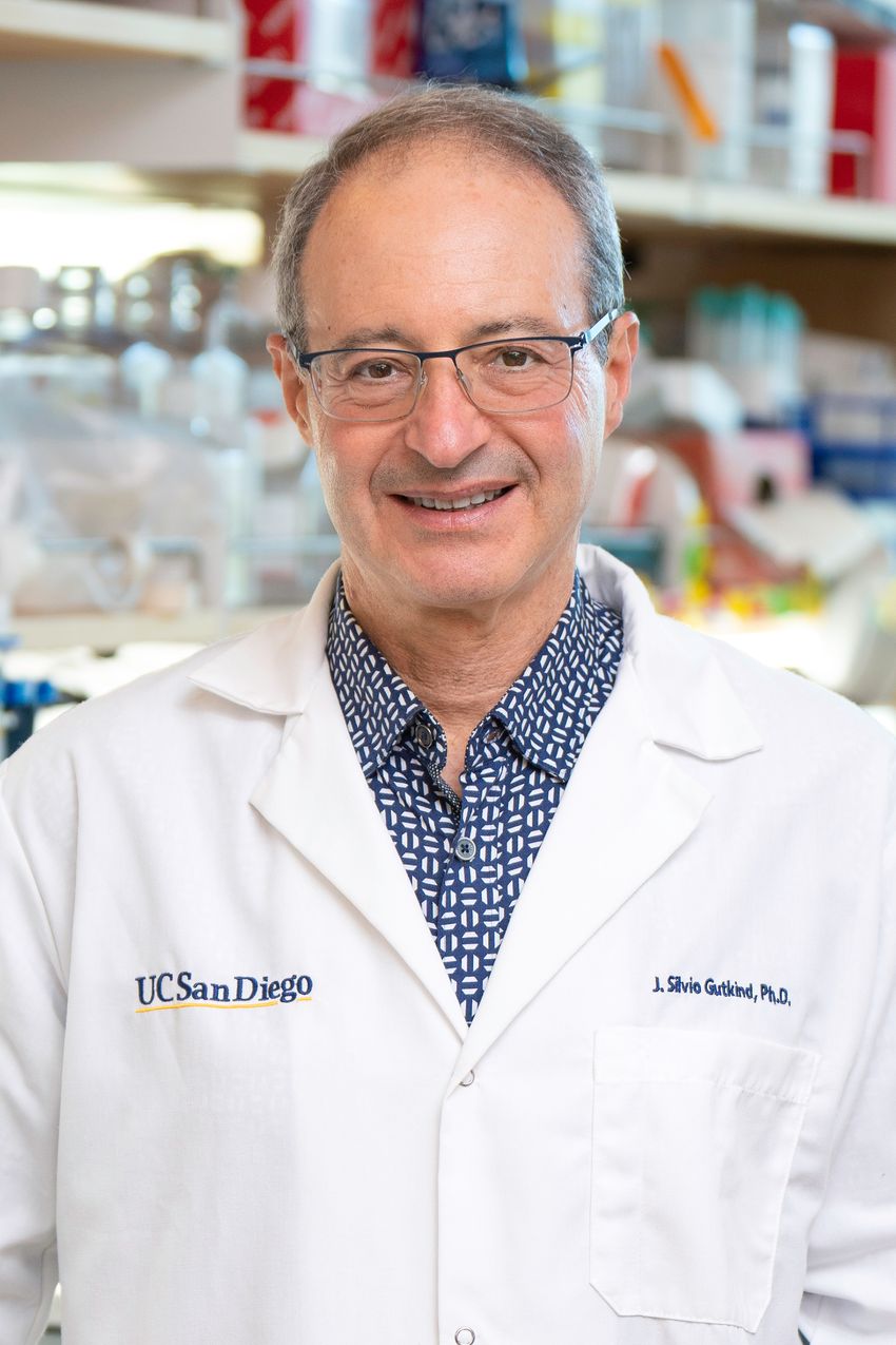 Jorge Silvio Gutkind in the laboratory. He’s wearing glasses, a blue shirt with whit patterns, and a lab coat embroidered with his name and “UCSanDiego.”
