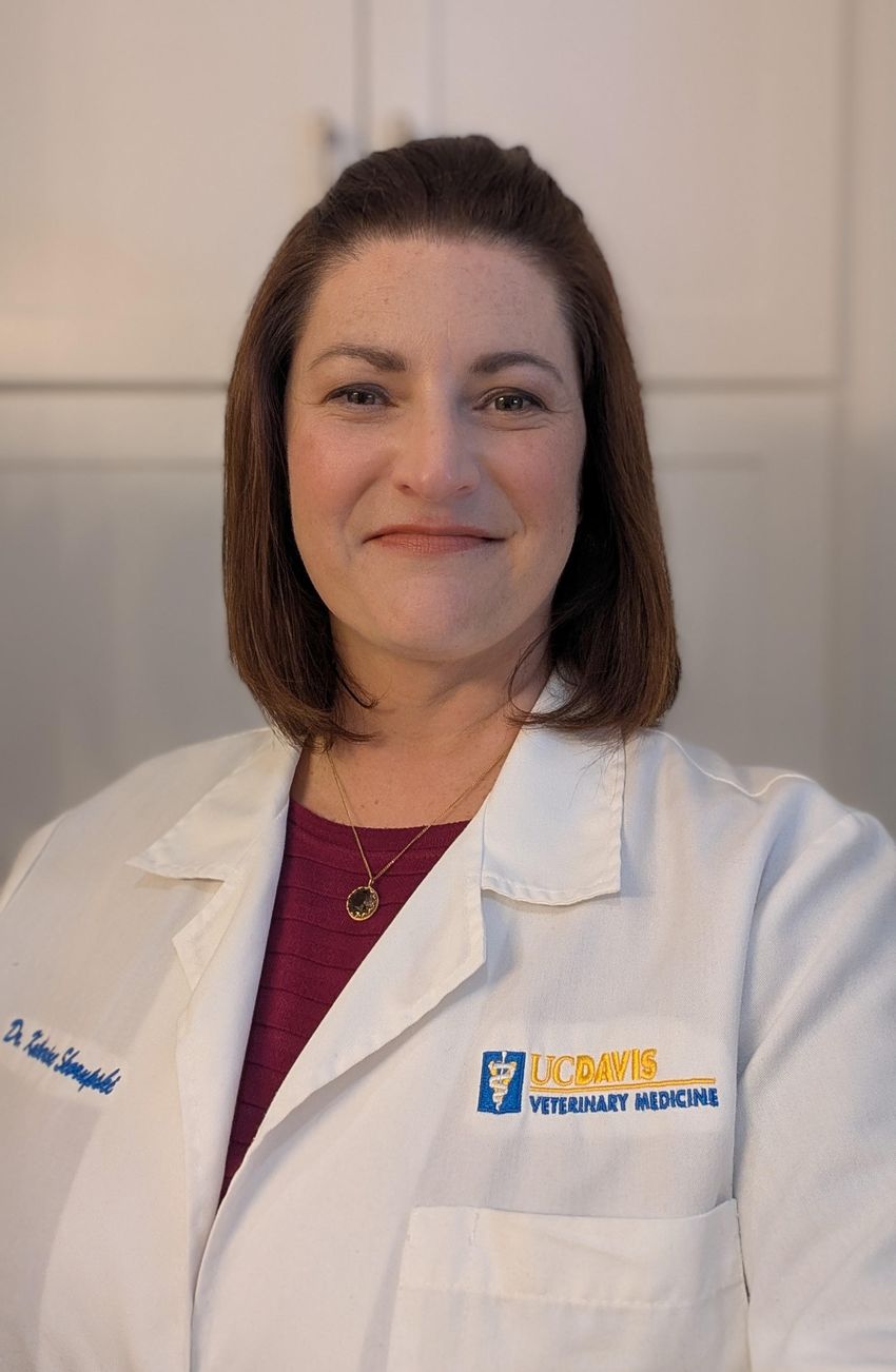 Katherine Skorupski poses in entrance of white cupboards. She’s carrying a purple shirt and a necklace beneath a white lab coat embroidered along with her title and “UC DAVIS VETERINARY MEDICINE
