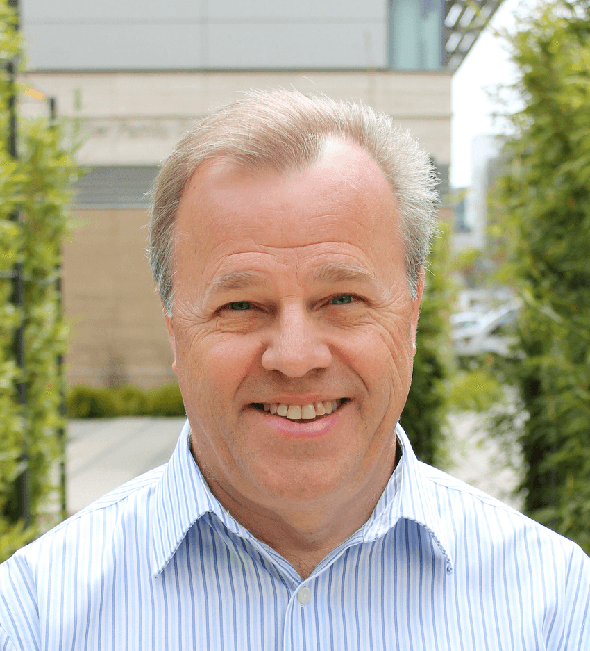 Daniel Johnson poses in front of a blurred background of a building with plants. He’s wearing a light blue shirt with vertical stripes.