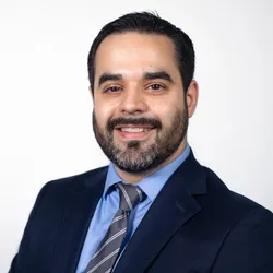 Photograph of Gregory Fonzo wearing a blue shirt, striped grey tie, and navy blue blazer.