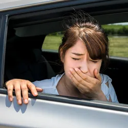 Image of a woman facing out of an open car window. She has one hand on the car and one hand covering her mouth. She appears carsick.