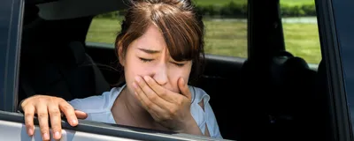 Image of a woman facing out of an open car window. She has one hand on the car and one hand covering her mouth. She appears carsick.