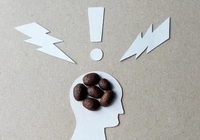 Image of a cutout silhouette of a person’s profile with six coffee beans clustered in their head. There are cutout shapes of two lightning bolts and an exclamation point above their head. Image of a cutout silhouette of a person’s profile with six coffee beans clustered in their head. There are cutout shapes of two lightning bolts and an exclamation point above their head.