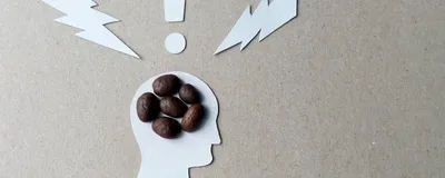 Image of a cutout silhouette of a person’s profile with six coffee beans clustered in their head. There are cutout shapes of two lightning bolts and an exclamation point above their head. 