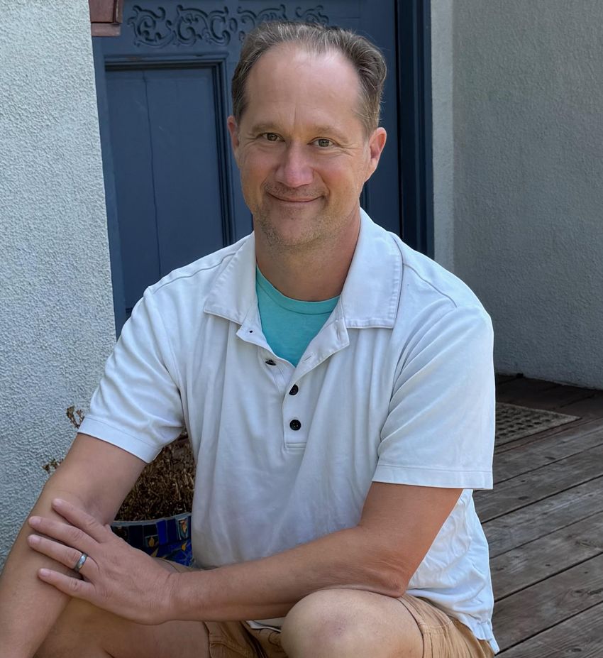 Van Savage sits on the front porch of a house with white walls, dark blue door, and wood-paneled floor. He’s wearing a white polo shirt with a blue tee underneath, brown short, and a wedding band on his left hand.