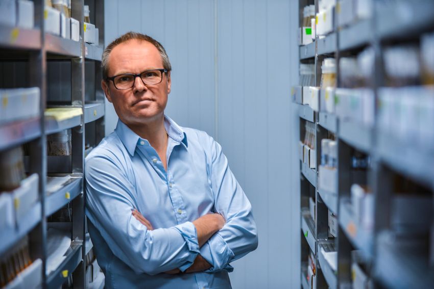 Gero Miesenböck, wearing a light blue shirt, poses with his arms folded. He was leaning against a shelving unit in front of a blue paneled background.