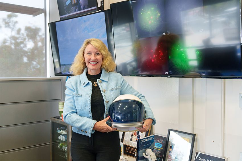 Catriona Jamieson, wearing a black top and pants as well as a light blue jacket, smiles at the camera while holding an astronaut helmet. She’s standing in a room with framed magazine clippings, a minifridge, and a large monitor displaying fluorescence microscopy images of cells in the background.