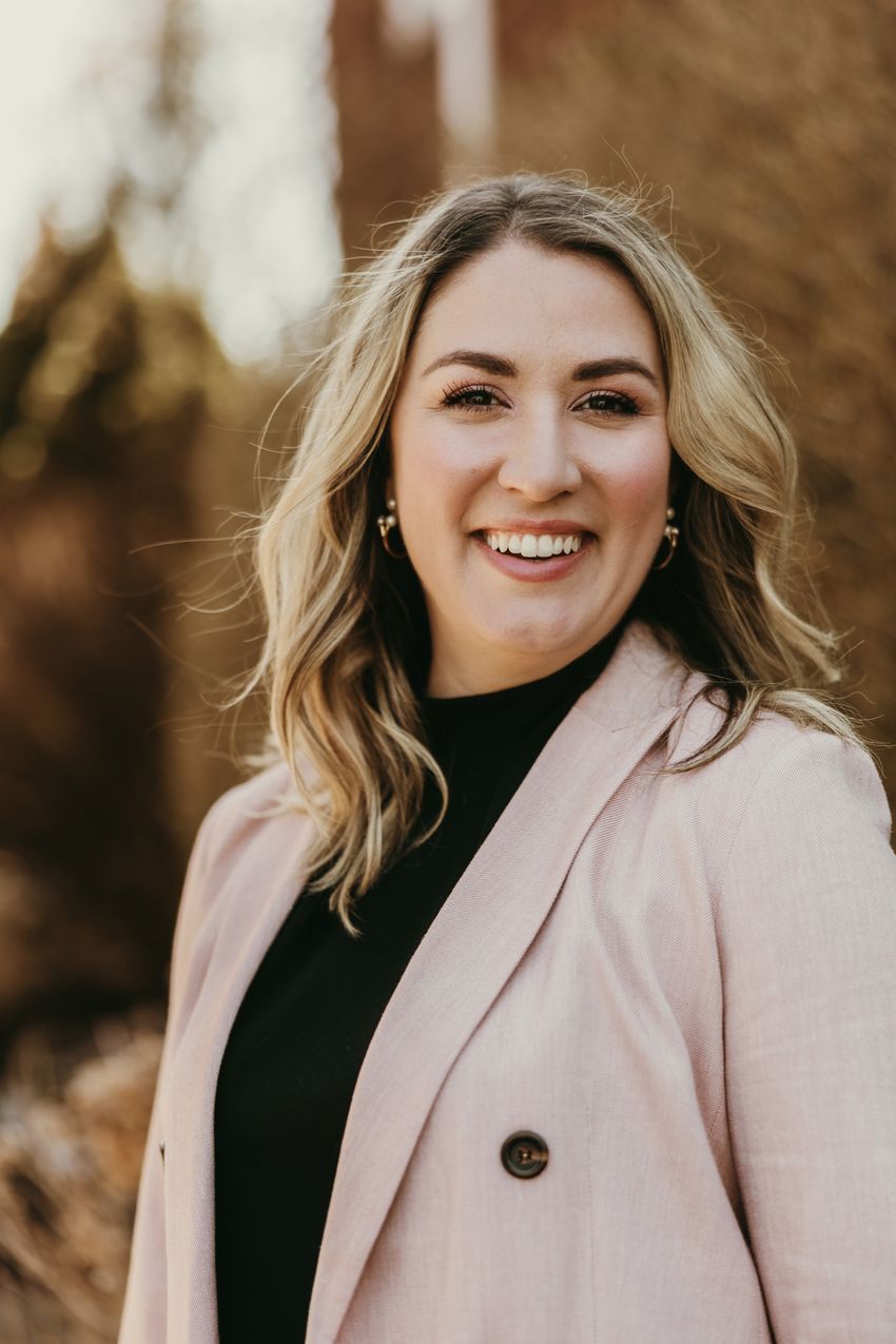 Abigail Overacre-Delgoffe, wearing a dark top and beige jacket with black button, smiles for the camera in front of a blurred background with dark, earthy tones.