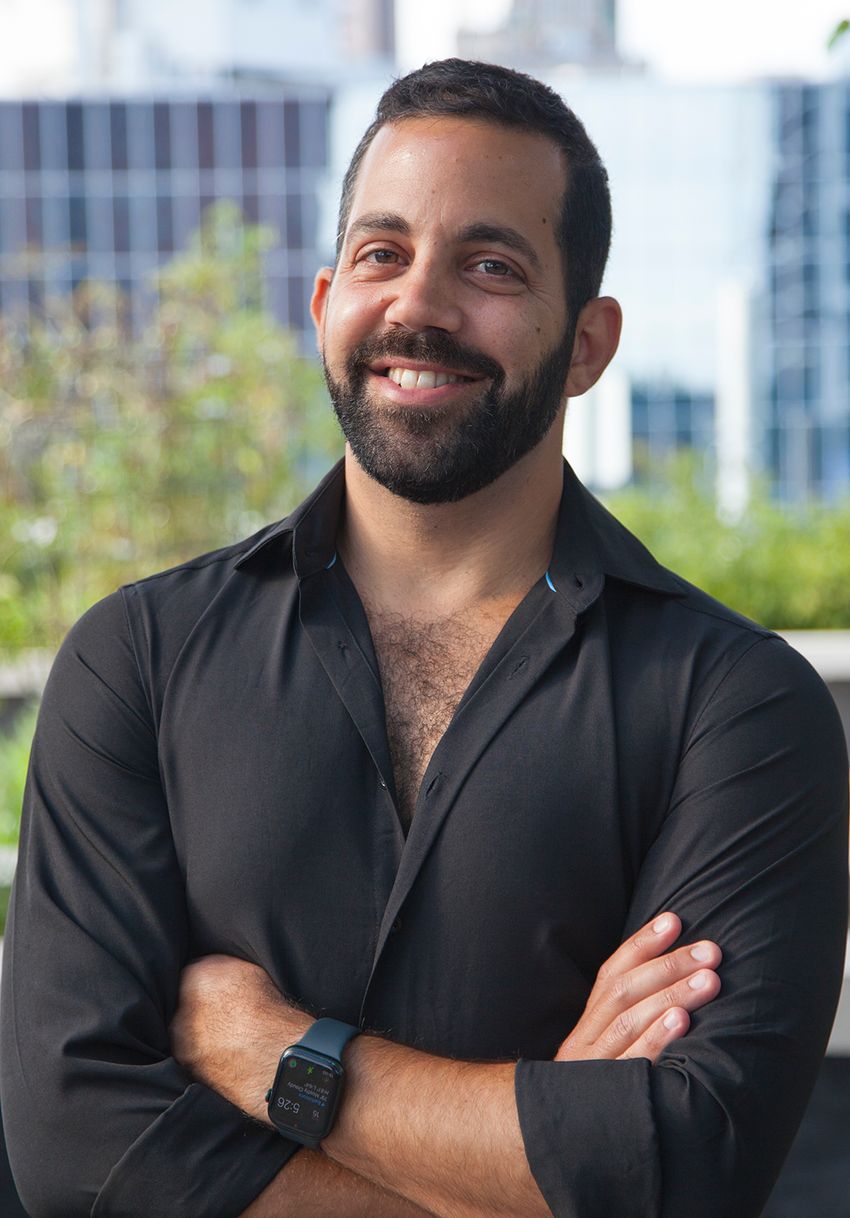Jotham Suez, wearing a black shirt and a black watch, poses with his arms crossed in front of a blurred background with glass buildings and trees.