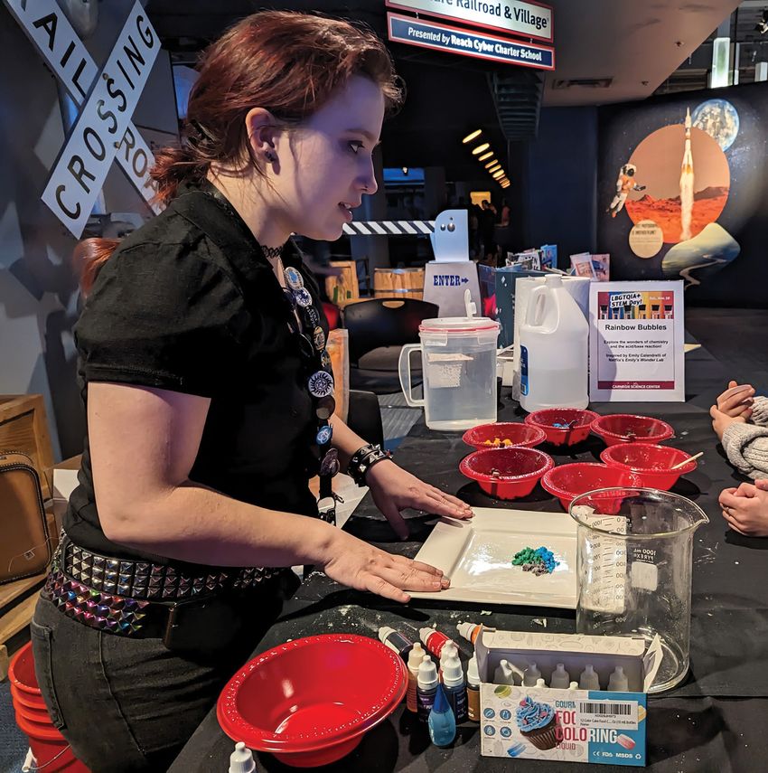 Image of Shelby Bradford standing at a table while performing a live demonstration of rainbow bubbles for museum attendees.