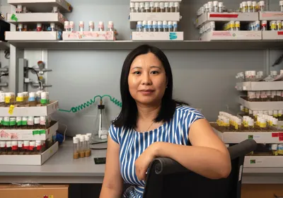 Image of Li Zhao. She smiles at the camera. She has one arm draped over the back of a chair. In the background, there are many labeled test tubes on a laboratory bench.