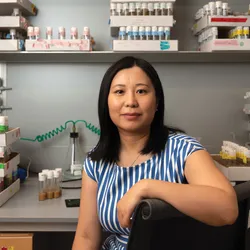 Image of Li Zhao. She smiles at the camera. She has one arm draped over the back of a chair. In the background, there are many labeled test tubes on a laboratory bench. Image of Li Zhao. She smiles at the camera. She has one arm draped over the back of a chair. In the background, there are many labeled test tubes on a laboratory bench.