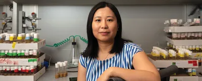 Image of Li Zhao. She smiles at the camera. She has one arm draped over the back of a chair. In the background, there are many labeled test tubes on a laboratory bench.