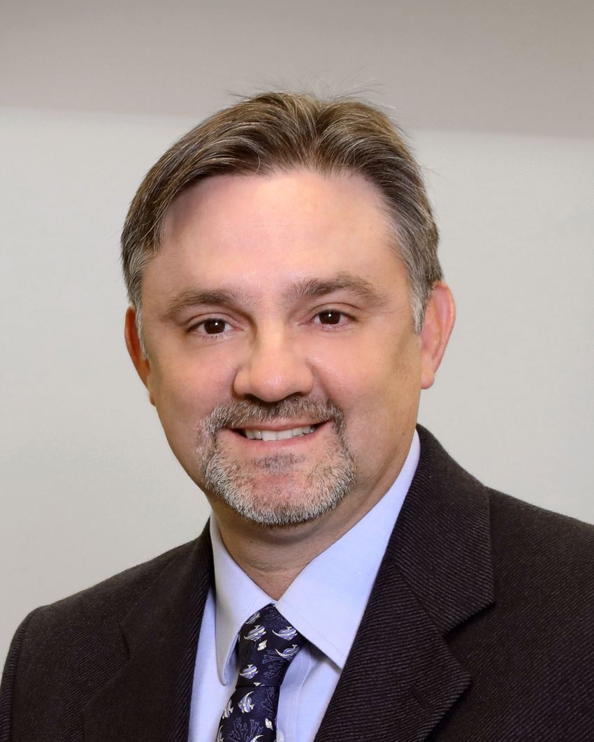 Jesús (Tito) González, wearing a light blue shirt, a patterned tie, and a black textured suit, smiles at the camera in front of a white wall.
