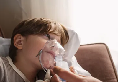 A child lies on their side on a sofa with a breathing apparatus covering their nose and mouth. Such equipment is commonly used for patients who suffer from cystic fibrosis, a once deadly disease that now has become manageable thanks to the triple combination drug Trikafta. Three researchers recently received this year’s Lasker award for their contribution to the development of this drug. 