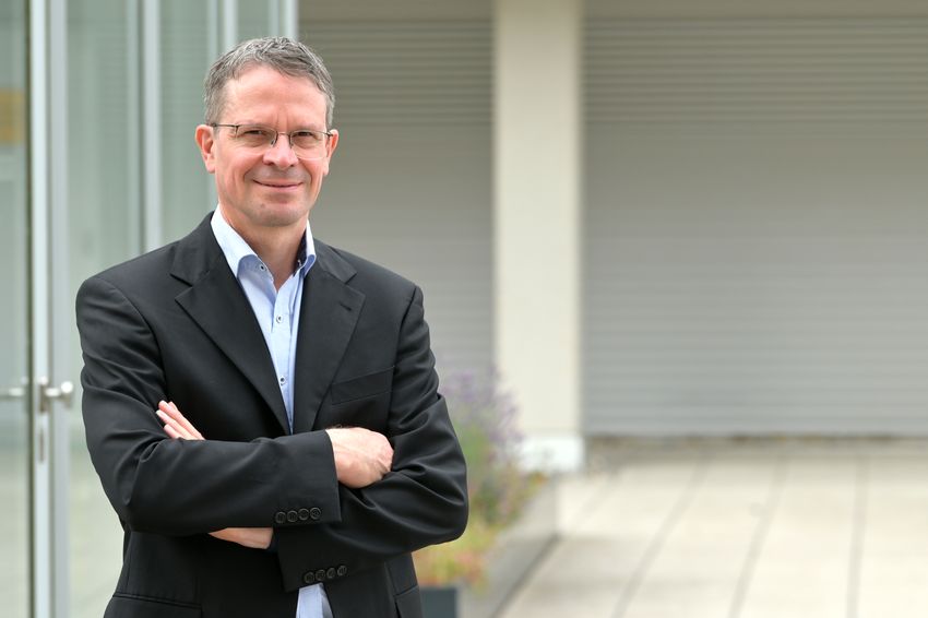 Photograph of Dirk Görlich, a biochemist at the Max Planck Institute for Multidisciplinary Science. He is wearing a black suit jacket over a light blue collared shirt and stands in an outdoor patio, with the background blurred.