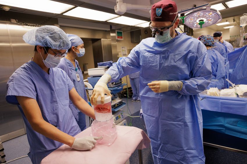 Surgeons stand in an operating theatre in blue gowns, gloves, and masks. One is holding a jar containing a pig kidney.