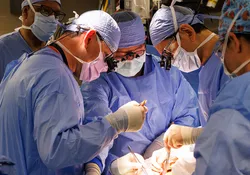 A team of surgeons wearing blue gowns, gloves, and masks are standing over a patient and operating. 