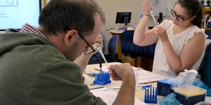 Photograph of two teachers, a woman in a white shirt with dark hair and glasses and a man with short dark hair in a green sweatshirt, at a table holding small plastic tubes and pipettes to perform DNA extraction. Photograph of two teachers, a woman in a white shirt with dark hair and glasses and a man with short dark hair in a green sweatshirt, at a table holding small plastic tubes and pipettes to perform DNA extraction.