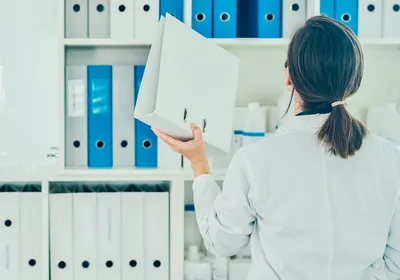 Photo of a woman in a lab coat placing a binder on a shelf. Photo of a woman in a lab coat placing a binder on a shelf.