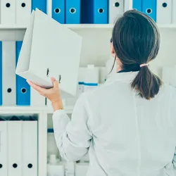 Photo of a woman in a lab coat placing a binder on a shelf. Photo of a woman in a lab coat placing a binder on a shelf.