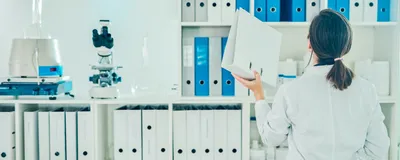 Photo of a woman in a lab coat placing a binder on a shelf. Photo of a woman in a lab coat placing a binder on a shelf.