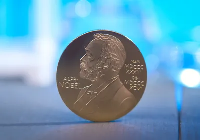 A gold medal depicting Alfred Nobel's side profile balances on a wooden surface in front of a blurred blue background. A gold medal depicting Alfred Nobel's side profile balances on a wooden surface in front of a blurred blue background.