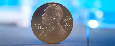 A gold medal depicting Alfred Nobel's side profile balances on a wooden surface in front of a blurred blue background. 