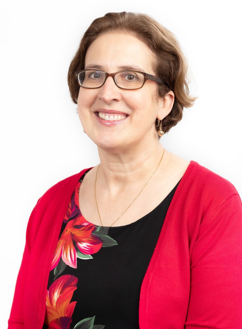 Jennifer Byrne, wearing a black top with red flowers, a red cardigan, and glasses, poses in front of a white background.