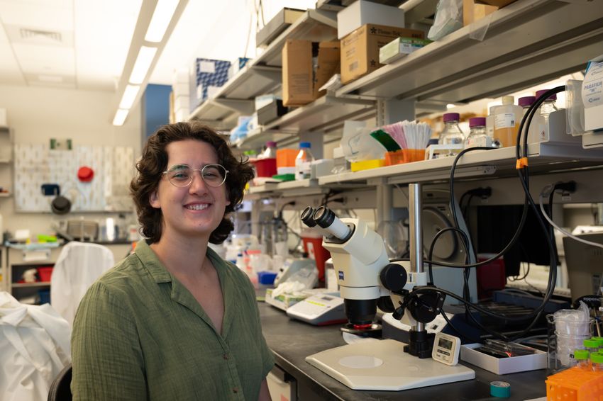 Photograph of Sarah Donofrio, a neuroscience graduate student at Baylor College of Medicine, sitting at a lab bench in front of a microscope. She has short dark hair, circle-framed glasses, and is wearing a green shirt and smiling at the camera.