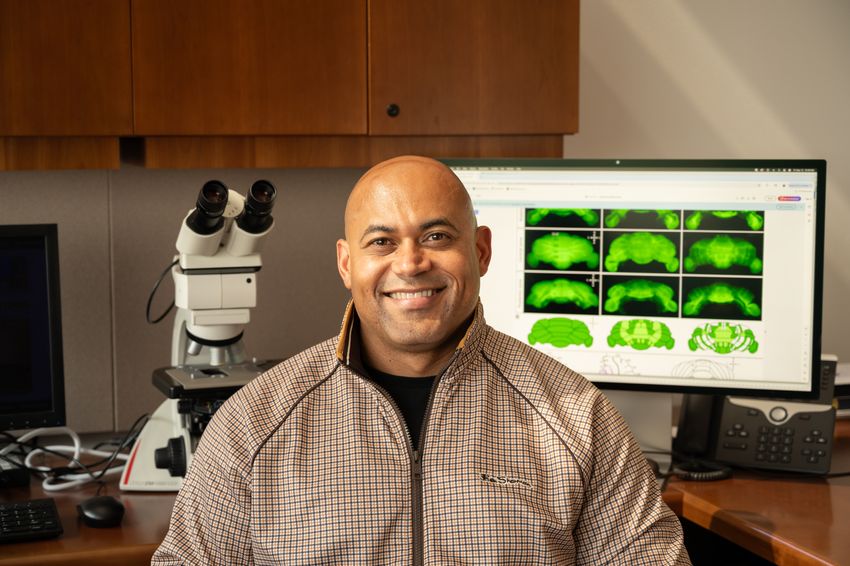Photograph of Roy Sillitoe, a neuroscientist at Baylor College of Medicine. Sillitoe sits in front of an office desk with a computer on the right and a microscope on the left of the image. He is wearing a zip-up jacket and is smiling at the camera.