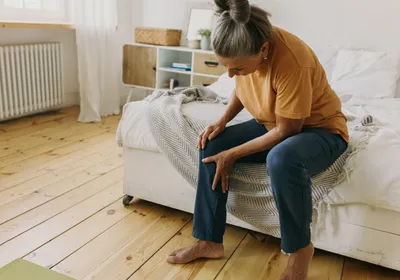 A woman with grey hair sits at the edge of her bed, bending down and holding her right knee. She seems to be in pain—joint pains are characteristic of patients with rheumatoid arthritis, though researchers recently found that their immune system may have become activated before their symptoms emerge. A woman with grey hair sits at the edge of her bed, bending down and holding her right knee. She seems to be in pain—joint pains are characteristic of patients with rheumatoid arthritis, though researchers recently found that their immune system may have become activated before their symptoms emerge.