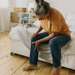A woman with grey hair sits at the edge of her bed, bending down and holding her right knee. She seems to be in pain—joint pains are characteristic of patients with rheumatoid arthritis, though researchers recently found that their immune system may have become activated before their symptoms emerge.