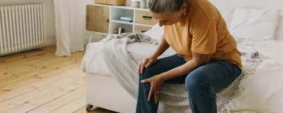 A woman with grey hair sits at the edge of her bed, bending down and holding her right knee. She seems to be in pain—joint pains are characteristic of patients with rheumatoid arthritis, though researchers recently found that their immune system may have become activated before their symptoms emerge. A woman with grey hair sits at the edge of her bed, bending down and holding her right knee. She seems to be in pain—joint pains are characteristic of patients with rheumatoid arthritis, though researchers recently found that their immune system may have become activated before their symptoms emerge.