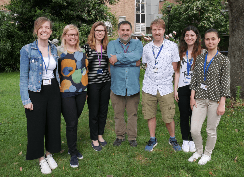 Members of Antal Rot’s lab stand together on grass in a semi-circle, facing the camera, in front of a building and some trees.