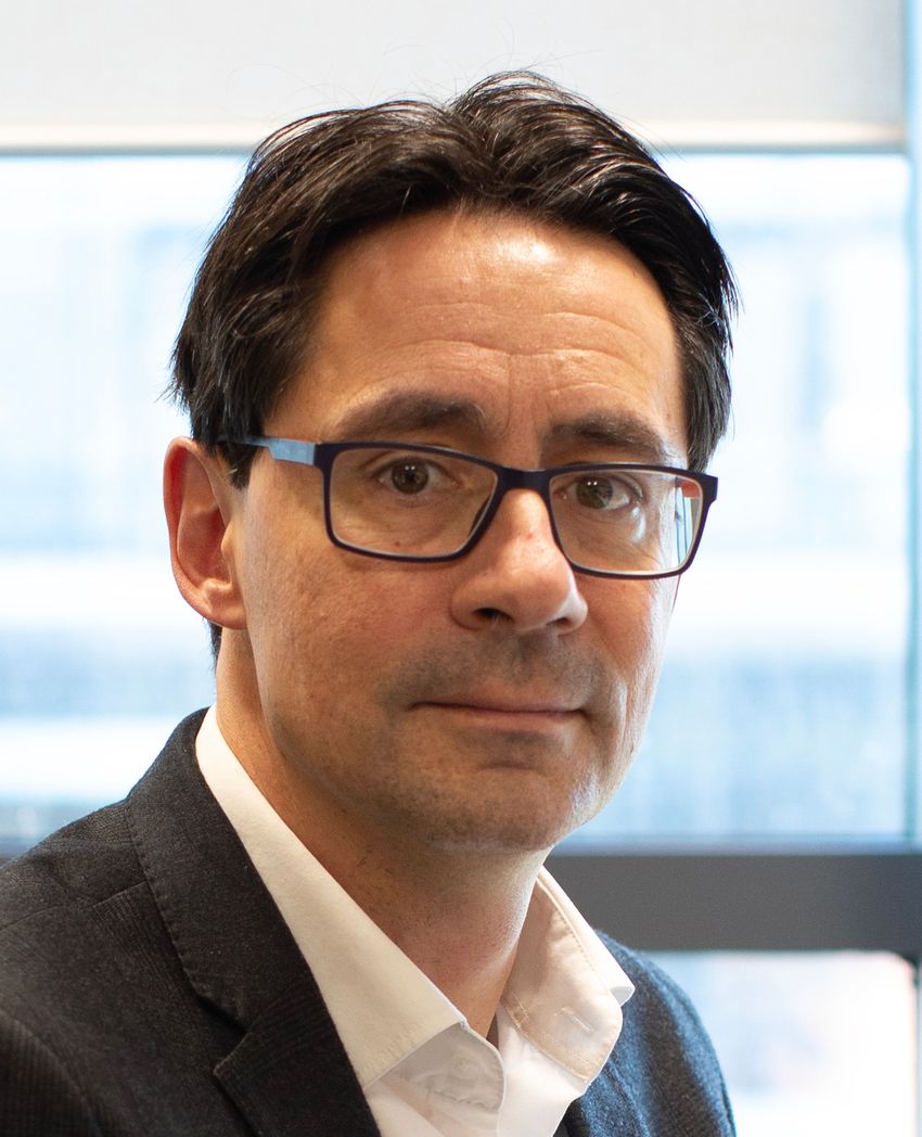 Profile shot of Cédric Ghevaert, wearing glasses, white shirt, and dark jacket, in front of a blurred background