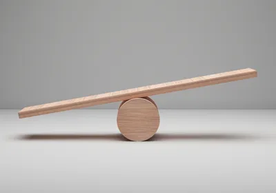 A photo of an unbalanced wooden plank resting on a round wooden ball against a neutral gray background.