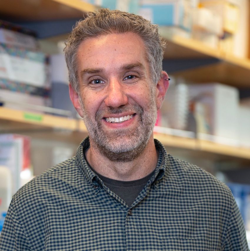James Fraser poses in front of a blurred background of a laboratory bench. He’s wearing a dark grey tee underneath a blue/green checkered shirt.