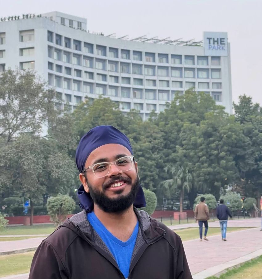 Onkar Gujral poses in a park, in front of a building called “THE PARK”. He’s wearing glasses, a blue T-shirt underneath a black jacket, and a dark blue headwrap.