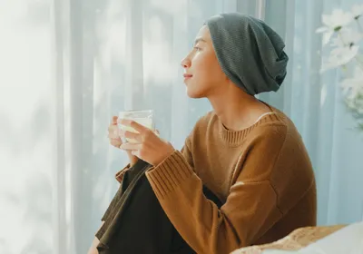 A woman wearing a large, loose headwear often worn by patients undergoing cancer chemotherapy, sits with her knees close to her body, holding a drink in a cup. She is dressed in a brown knit sweater and dark brown pants, gazing into the distance, signifying the thoughtful ways that researchers have revolutionized cancer therapeutics. A woman wearing a large, loose headwear often worn by patients undergoing cancer chemotherapy, sits with her knees close to her body, holding a drink in a cup. She is dressed in a brown knit sweater and dark brown pants, gazing into the distance, signifying the thoughtful ways that researchers have revolutionized cancer therapeutics.