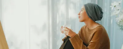 A woman wearing a large, loose headwear often worn by patients undergoing cancer chemotherapy, sits with her knees close to her body, holding a drink in a cup. She is dressed in a brown knit sweater and dark brown pants, gazing into the distance, signifying the thoughtful ways that researchers have revolutionized cancer therapeutics. 