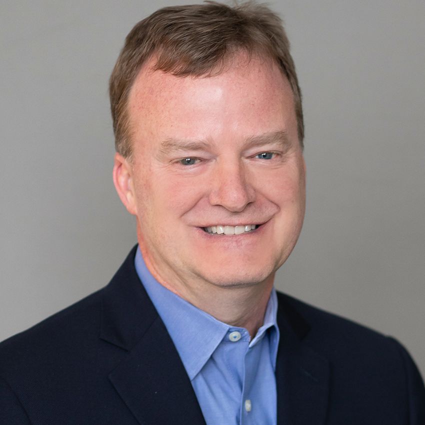 Paul Limburg wears a dark suit jacket and blue shirt in a headshot taking against a grey background.