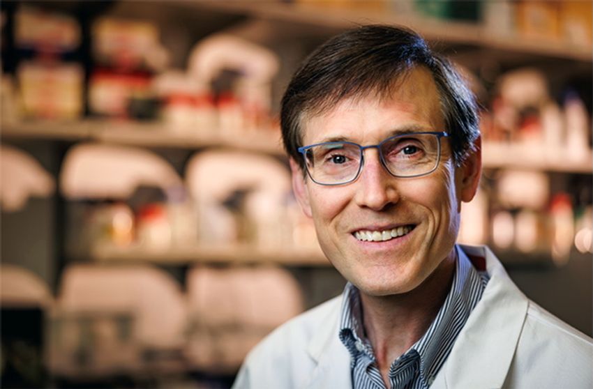 The gastroenterologist, William Grady wears glasses, a striped shirt, and a white lab coat in a headshot taking in a biology laboratory.