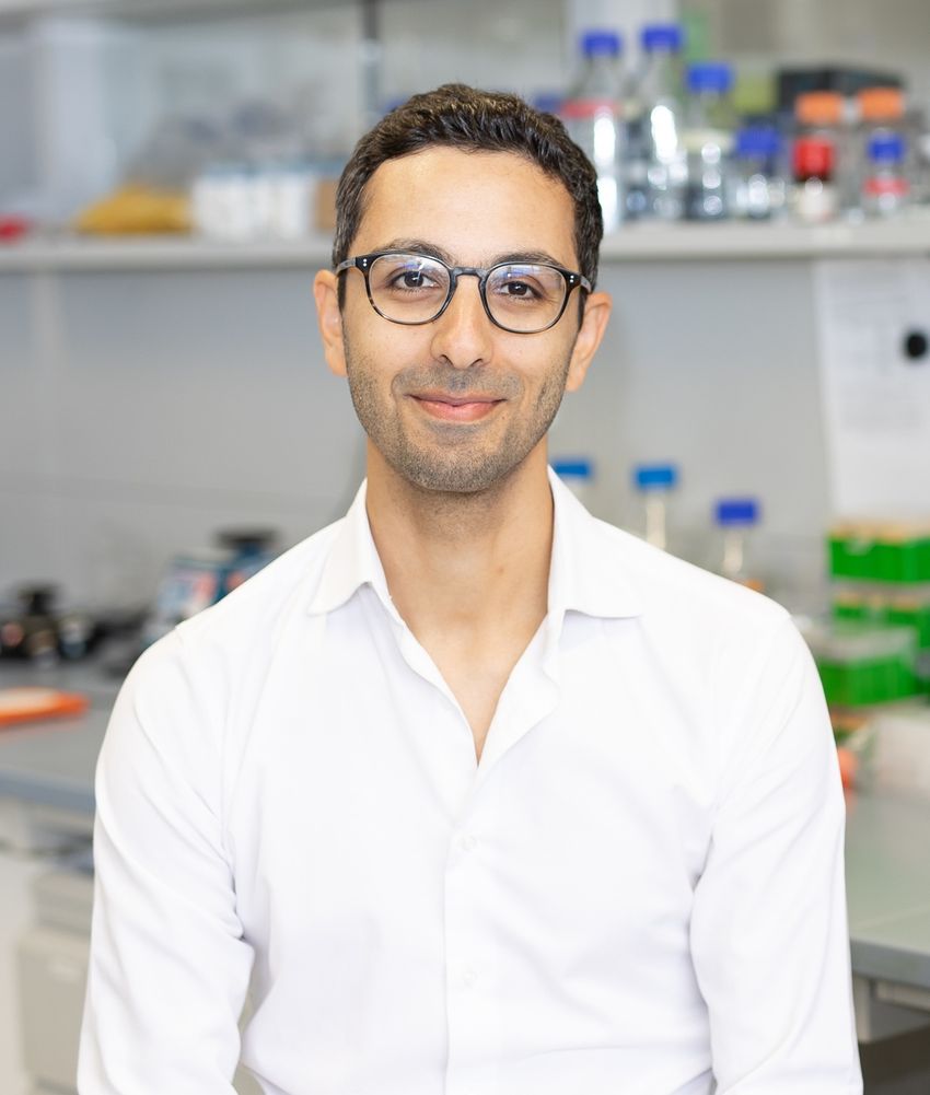 Scientist Thierry Nordmann, who studies skin diseases using deep visual proteomics, is smiling and wearing a white button-down shirt and black-framed glasses. In the background there are laboratory shelves of solutions in glass bottles. Scientist Thierry Nordmann, who studies skin diseases using deep visual proteomics, is smiling and wearing a white button-down shirt and black-framed glasses. In the background there are laboratory shelves of solutions in glass bottles.
