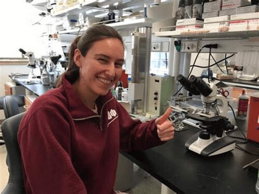 Photograph of Emma Dauster, today a senior script writer for Complexly: SciShow, sitting at a laboratory bench in front of a microscope. Dauster is wearing a maroon sweatshirt and has her brown hair tied back in a ponytail. She is smiling and giving a thumbs up. Photograph of Emma Dauster, today a senior script writer for Complexly: SciShow, sitting at a laboratory bench in front of a microscope. Dauster is wearing a maroon sweatshirt and has her brown hair tied back in a ponytail. She is smiling and giving a thumbs up.