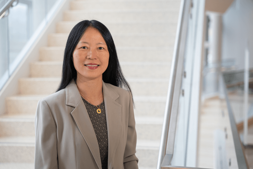 Zhiping Weng, a computational biologist, wears a light grey suit jacket and stands in front of a staircase in a headshot photo.